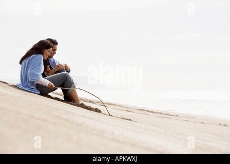 Coppia giovane seduto sulla spiaggia, donna disegno in sabbia con bastone Foto Stock