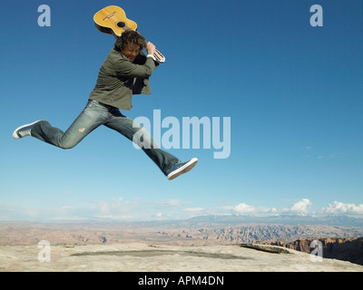 Metà uomo adulto con la chitarra acustica jumping, strati di roccia in background Foto Stock