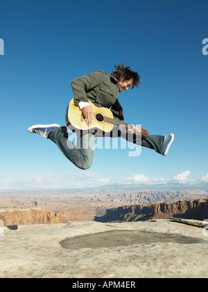 Metà uomo adulto jumping e suonare la chitarra acustica, strati di roccia in background Foto Stock