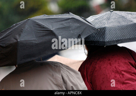 Trainare la gente camminare sotto la pioggia indossando indumenti impermeabilizza e ombrelli azienda sopra le loro teste in misere condizioni atmosferiche umide Foto Stock