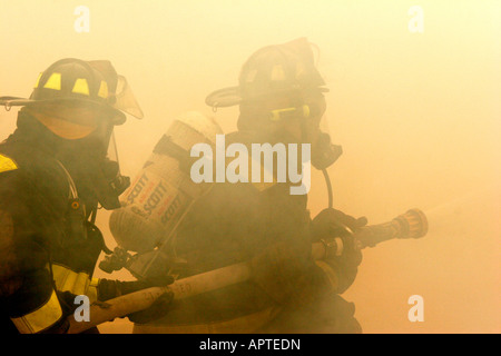 Due vigili del fuoco su un hoseline la spruzzatura di acqua in un incendio circondato dal fumo di un giallo errie scena di emergenza Foto Stock