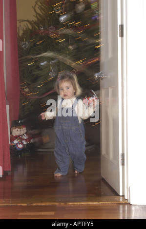 A due anni di età ragazza caucasica camminando attraverso una porta tenendo un giocattolo con un tradizionale inglese albero di Natale dietro di lei, Foto Stock