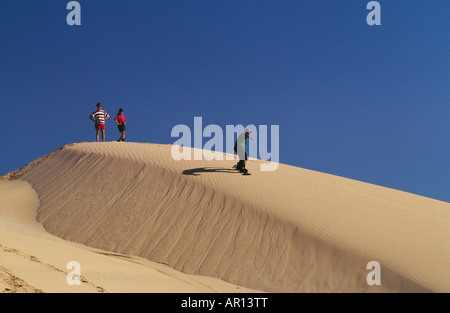 Un uomo scorre sulle dune di sabbia con uno scarpone da snowboard come un paio di guardare a lui Foto Stock