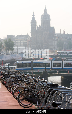 HOLLAND AMSTERDAM vista di biciclette parcheggiate nel multipiano Bike Park con il tram viaggiano PASSATO E CHIESA IN BACKGROUND Foto Stock