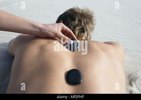 Giovane donna immissione black hot stones sull'uomo torna sdraiato sulla spiaggia Foto Stock