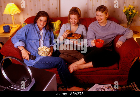 Le coinquiline, in stile anni '1990, pizze e popcorn, condividono un divano e guardano una piccola TV portatile nel loro soggiorno condiviso in affitto Foto Stock