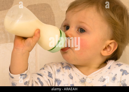 Un bambino di un anno che tiene e beve dal suo biberon del latte vestito per il letto Foto Stock