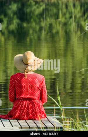 Backview di una donna in abito rosso con il cappello per il sole seduti sulla passerella di legno in un lago Foto Stock