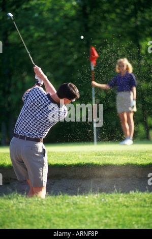 Golfista maschio chip dalla trappola di sabbia sul verde, femmina golfista si erge dal foro tiene la bandiera, British Columbia, Canada. Foto Stock