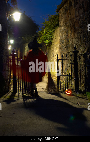 Silhouette di una giovane donna che indossa un mantello rosso e una strega hat a piedi attraverso un piccolo cancello di ferro in un vicolo solitario di notte Foto Stock