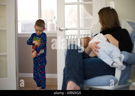 Madre allattamento neonato a casa mentre quattro anni di fratello si erge nelle vicinanze Foto Stock