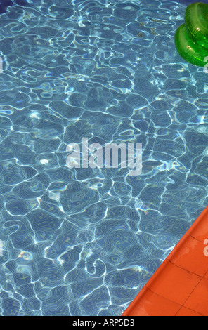 Abstract di piscina blu con luce solare che gioca sulla superficie. Parte di verde gonfiabile nell'angolo in alto a destra e piastrelle rosse in basso a destra. Foto Stock