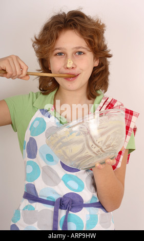 Ragazza adolescente rendendo la cottura la cottura di torte con pinafore e intestino leccare il cucchiaio di legno Spatola Foto Stock