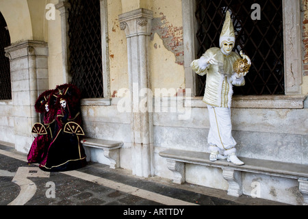 Un uomo in Costume di Carnevale e la maschera in equilibrio su un banco di pietra di essere guardato da due donna vestita simili Foto Stock