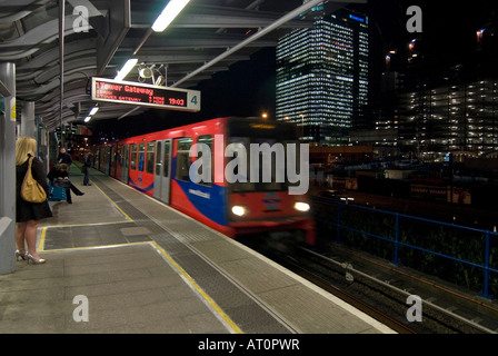 In orizzontale ampia angolazione di una Docklands Light Railway in treno arrivando alla stazione di pioppo con persone in attesa sulla piattaforma di notte Foto Stock