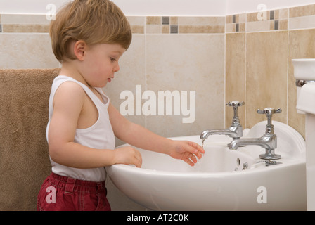 bambino di ventidue mesi lavando e usando i rubinetti dell'acqua del rubinetto nel lavandino in bagno, vestito con giubbotto bianco, singolo Foto Stock