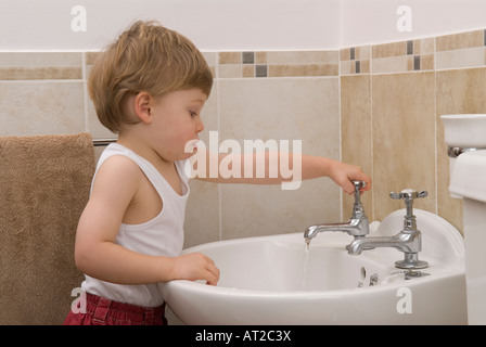 bambino di ventidue mesi lavando e usando i rubinetti dell'acqua del rubinetto nel lavandino in bagno, vestito con giubbotto bianco, singolo Foto Stock