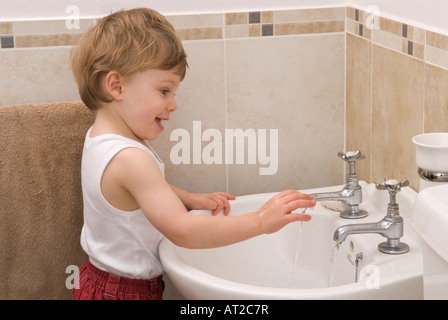 bambino di ventidue mesi lavando e usando i rubinetti dell'acqua del rubinetto nel lavandino in bagno, vestito con giubbotto bianco, singolo Foto Stock