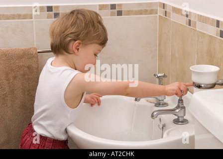 bambino di ventidue mesi lavando e usando i rubinetti dell'acqua del rubinetto nel lavandino in bagno, vestito con giubbotto bianco, singolo Foto Stock