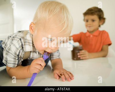 Due ragazzi (4-5), (12-24 mesi), ragazzo leccare il cioccolato dal cucchiaio, ritratto Foto Stock