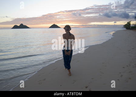 Un alto giovane donna cammina lungo la splendida spiaggia di Lanikai Beach all'alba con le isole Mokulua nella distanza. Foto Stock
