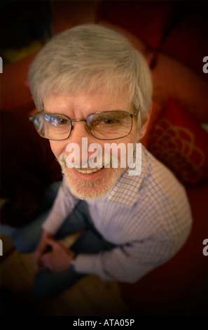Uomo anziano con i capelli grigi e gli occhiali seduto su una sedia guardando in alto nella fotocamera a sorridere Foto Stock