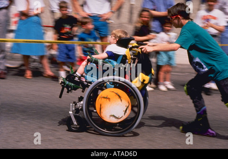 Bambino con handicap in carrozzella essendo spinto dal fratello su pattini in una sfilata Foto Stock