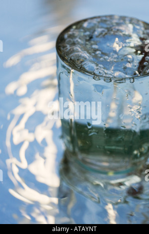 Versare acqua in un bicchiere che si erge su uno specchio che riflette il cielo blu Foto Stock