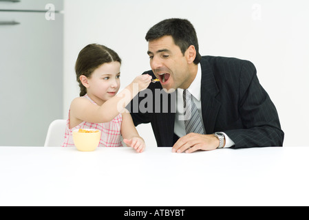 Bambina alimentazione di cereali al suo padre, sia la seduta a tavola Foto Stock