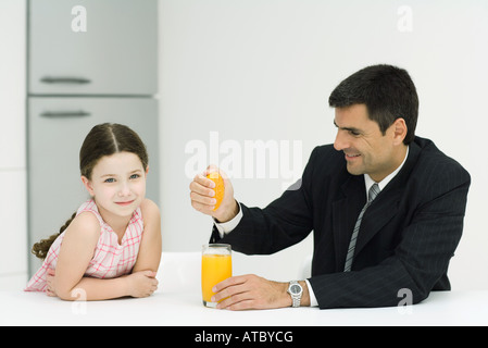 Padre e figlia seduti insieme a tavola, l'uomo la spremitura di succhi d' arancia in vetro, ragazza sorridente in telecamera Foto Stock