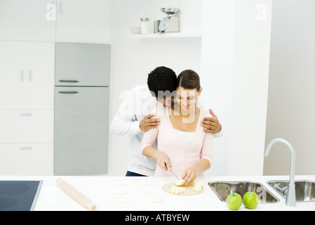 Matura in piedi insieme in cucina, donna il taglio di mele, uomo testa pendente sulla sua spalla Foto Stock