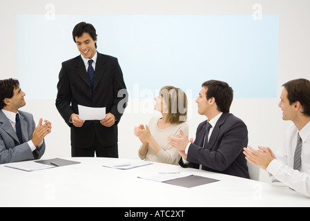 Imprenditore in piedi, tenendo documento, colleghi seduti battimani, sorridente Foto Stock