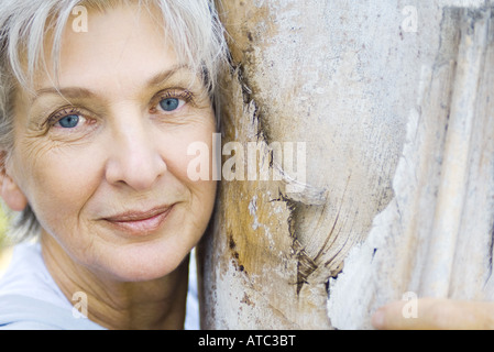Senior donna appoggiata contro il tronco di albero, sorridente in telecamera, ritratto Foto Stock