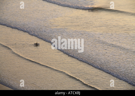 Un singolo surfer esce fuori verso il mare nella luce della sera portando la sua pensione Foto Stock
