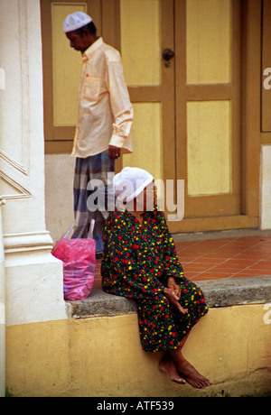 Arabo shoeless donna seduta outdoor per una moschea locale come un uomo passa da dietro il suo Singapore Sud-est asiatico Foto Stock