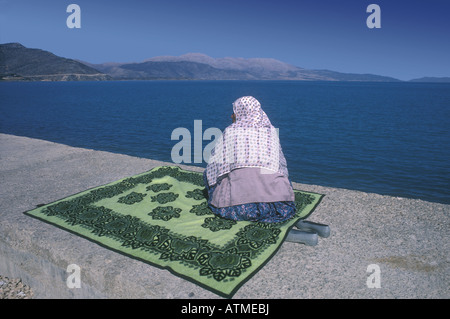 Donna che indossa hijab rosa si siede su un tappeto turco verde a Kale accanto al vuoto blu del lago Egirdir, Turchia - Türkiye Foto Stock