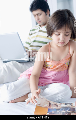 Close-up di una ragazza leggendo una rivista con suo padre a lavorare su un laptop dietro di lei Foto Stock