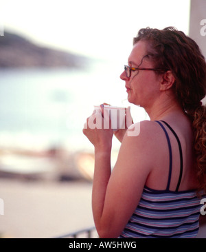 Giovane donna in vacanza avente una tazza di caffè sul balcone che si affaccia sulla spiaggia Foto Stock