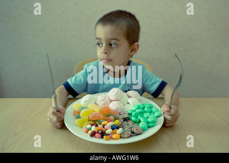 Little Boy tenendo il coltello e forchetta guarda un piatto pieno di caramelle che somigliano a un pasto Foto Stock