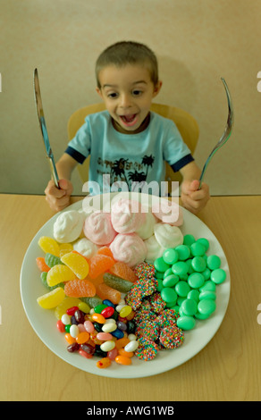 Little Boy tenendo il coltello e forchetta guarda un piatto pieno di caramelle che somigliano a un pasto Foto Stock