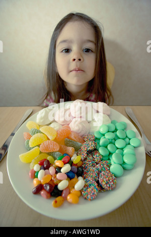 Bambina a tavola con coltello e forchetta e un piatto pieno di caramelle che somigliano a un pasto Foto Stock