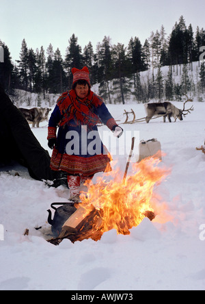 Finlandia, Saami donna in piedi accanto al fuoco, slitta trainata da renne in background Foto Stock