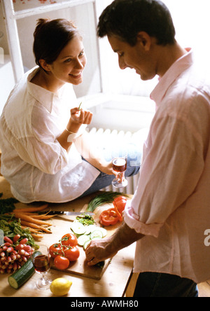 L'uomo tritare verdure sul tavolo, donna seduta sul tavolo, tenendo il vetro Foto Stock