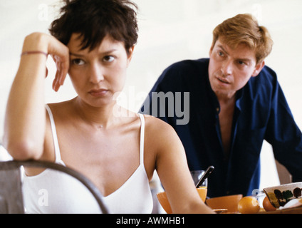 Donna seduta, gomito sul retro della sedia, mano contro la testa, uomo proteso verso di lei in background, vista frontale Foto Stock