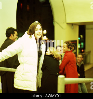 Giovane donna in cappotto sorridente alla fotocamera, gruppo di giovani in background Foto Stock