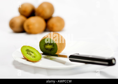 Single kiwi tagliata aperta su una piastra con coltello, più kiwi accatastati sul retro Foto Stock