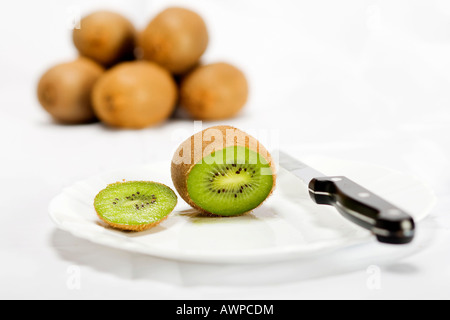 Single kiwi tagliata aperta su una piastra con coltello, più kiwi accatastati sul retro Foto Stock