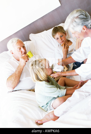 I nonni e i bambini a mangiare la prima colazione sul letto, donna aiutando bambina bere dalla tazza Foto Stock