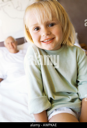 Bambina sul letto, sorridente alla fotocamera, nonno in background Foto Stock