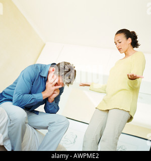 Donna in piedi, parlando e gesticolando per l'uomo, l'uomo seduto con le mani sul viso Foto Stock
